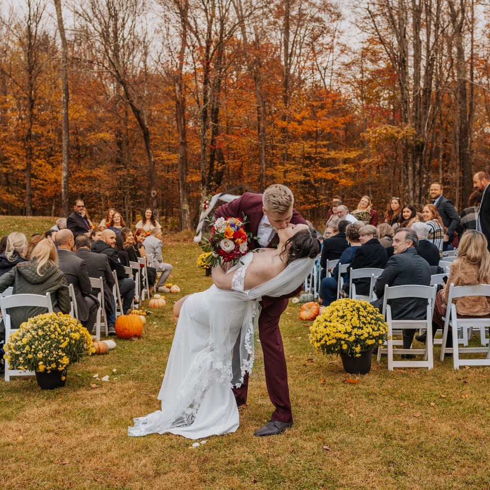 Groom in a maroon suit dips and kisses his bride in a white dress at an outdoor autumn wedding.