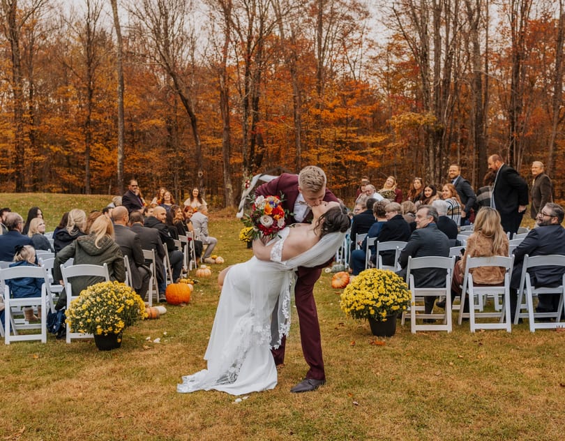 Groom in a maroon suit dips and kisses his bride in a white dress at an outdoor autumn wedding.